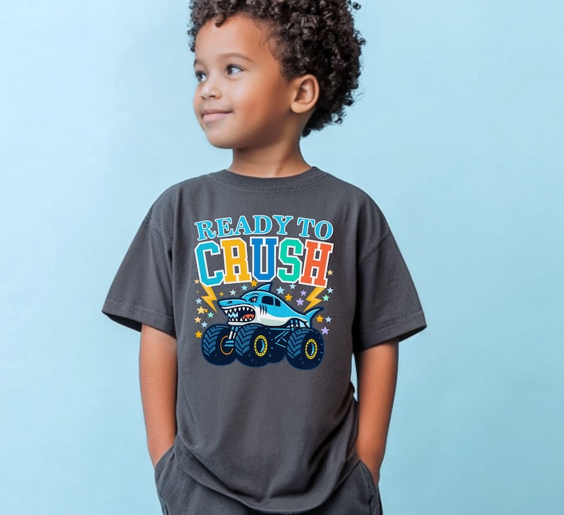 May include: Dark gray t-shirt with the text "READY TO CRUSH" above a cartoon shark monster truck. The shark is light blue with a white belly and a toothy grin. The truck has large black tires with yellow rims.