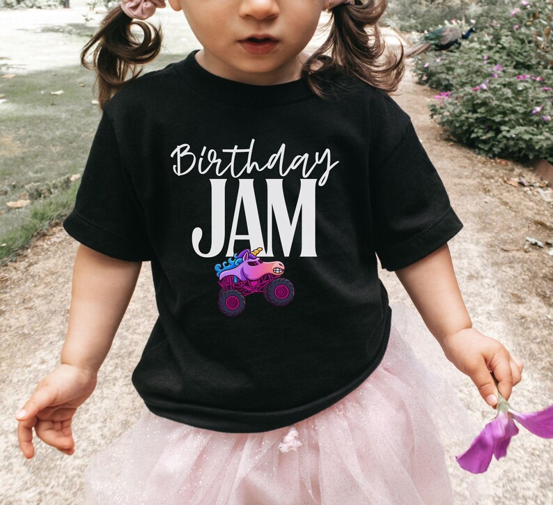 May include: Black t-shirt with the words "Birthday JAM" in white script and block letters. A colorful graphic of a unicorn monster truck is below the text. The shirt is worn by a child.