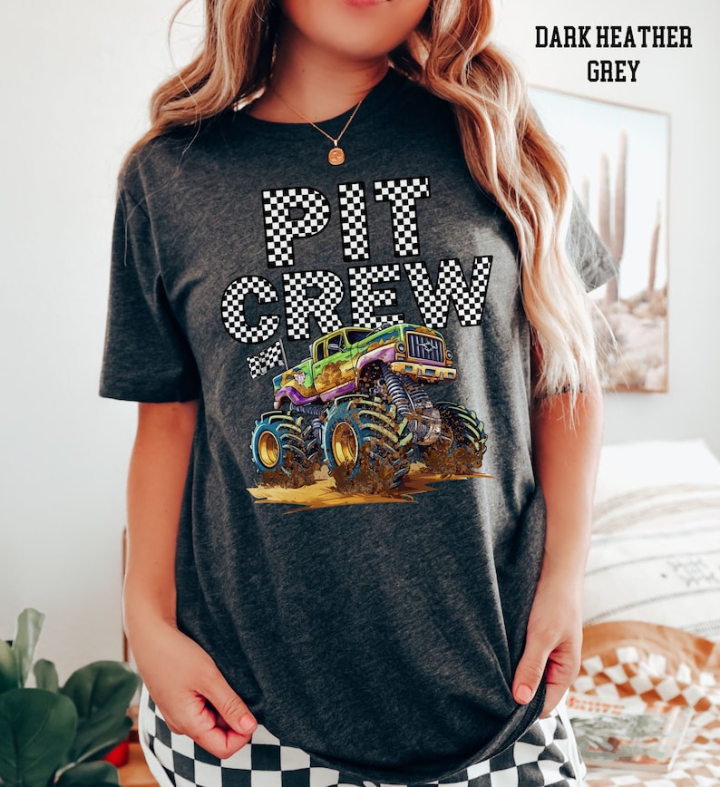 May include: Dark heather grey t-shirt with the words "PIT CREW" in a checkered pattern. A colorful monster truck graphic is featured below the text. The shirt is a comfortable, casual style.