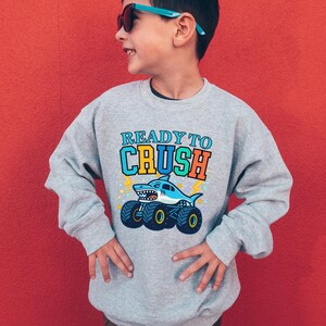 Pu&ograve; includere: Una felpa grigia con la scritta "READY TO CRUSH" sopra un cartone animato di uno squalo che guida un monster truck. Lo squalo &egrave; blu e bianco, e il camion ha grandi ruote nere e gialle. Lo sfondo &egrave; rosso pieno.