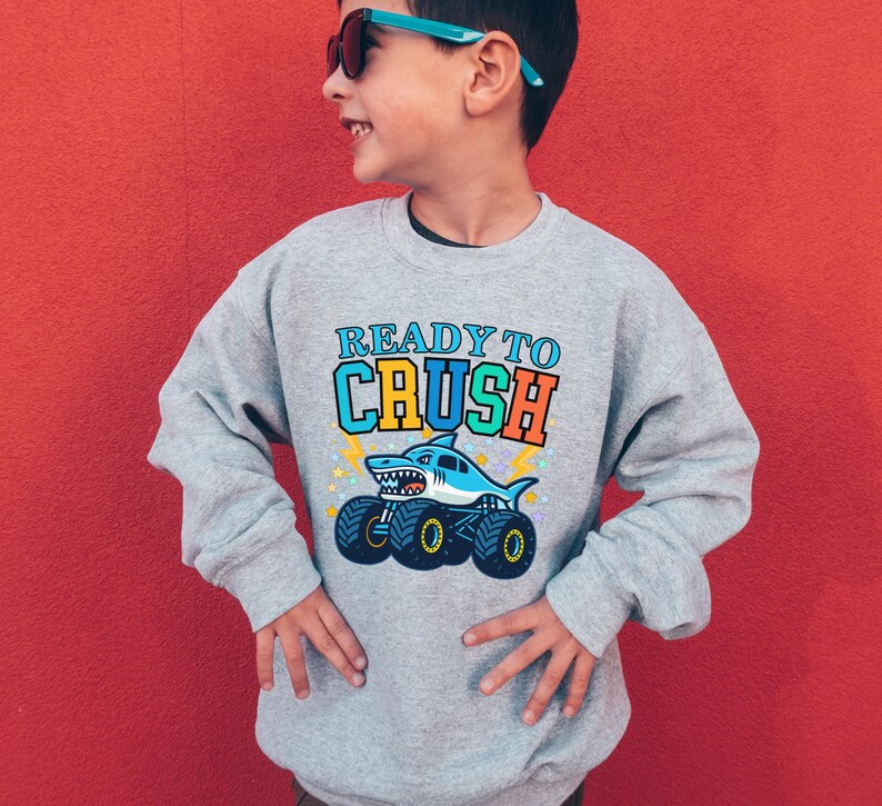 Pu&ograve; includere: Una felpa grigia con la scritta "READY TO CRUSH" sopra un cartone animato di uno squalo che guida un monster truck. Lo squalo &egrave; blu e bianco, e il camion ha grandi ruote nere e gialle. Lo sfondo &egrave; rosso pieno.