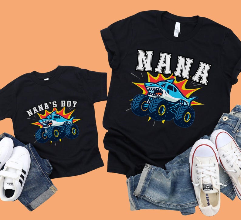 May include: Black t-shirts with matching shark monster truck graphics. One shirt says "NANA" and the other says "NANA'S BOY". The shark is light blue with a white belly and a menacing grin. The monster truck has blue tires with yellow accents.