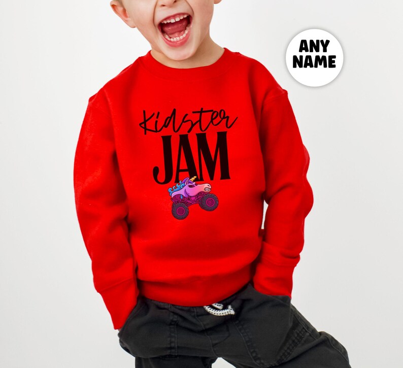 May include: A red long-sleeved sweatshirt with the words "Kidster JAM" in black script and block letters. A colorful monster truck graphic is below the text. The sweatshirt is designed for children.