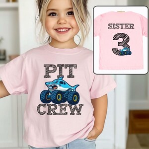 Op de afbeelding: Een lichtroze t-shirt met de tekst "PIT CREW" en een monster truck-ontwerp met haaienthema. Het shirt toont ook een kleinere afbeelding van een roze t-shirt met de tekst "SISTER 3" en een monstertruck.