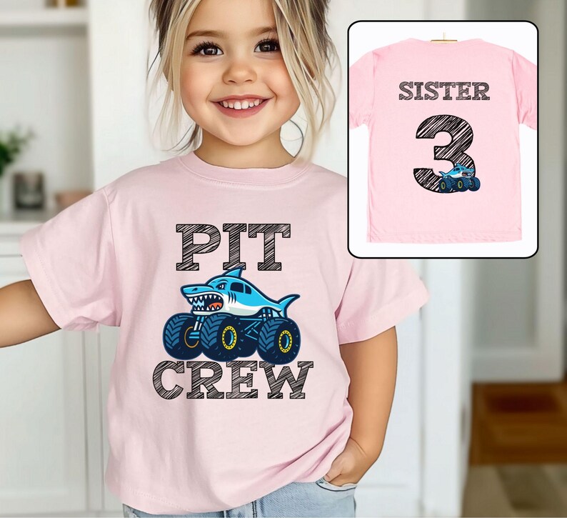 Op de afbeelding: Een lichtroze t-shirt met de tekst "PIT CREW" en een monster truck-ontwerp met haaienthema. Het shirt toont ook een kleinere afbeelding van een roze t-shirt met de tekst "SISTER 3" en een monstertruck.