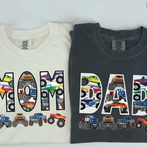 May include: Two t-shirts, one ivory and one dark gray, with the words "MOM" and "DAD" spelled out in large white letters. The letters are filled with a pattern of colorful monster trucks. The shirts are a matching set.