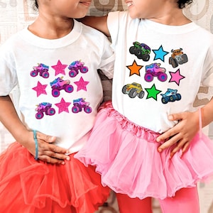 May include: Two children wearing white t-shirts with colorful graphics. One shirt features pink stars and unicorn monster trucks, the other has monster trucks and stars in various colors. Both are paired with pink tutus.