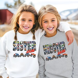 May include: Two children wearing gray and white sweatshirts with a monster truck design. The text on the sweatshirts reads "Kidster Jam" and "Sister Jam".