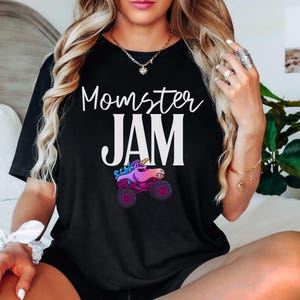 May include: Black t-shirt with the words "Momster JAM" in white script and block letters. A colorful cartoon monster truck with a unicorn horn is below the text. The shirt is a casual style.