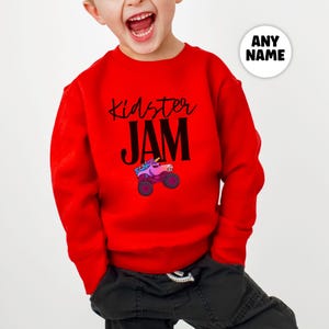 May include: A red long-sleeved sweatshirt with the words "Kidster JAM" in black script and block letters. A colorful monster truck graphic is below the text. The sweatshirt is designed for children.