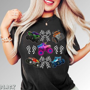 May include: Black t-shirt featuring colorful monster truck graphics with checkered bows. The trucks include a green monster truck, an orange truck, a unicorn truck, a red truck, and a shark truck.
