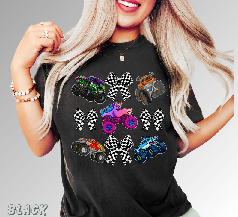 May include: Black t-shirt featuring colorful monster truck graphics with checkered bows. The trucks include a green monster truck, an orange truck, a unicorn truck, a red truck, and a shark truck.