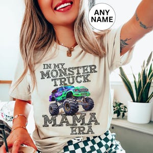 model is wearing a natural colored t-shirt with black chalk art letters saying " In My Monster Truck Mama Era" with a green and purple truck. Personalized Name shirt. Family Matching. Monster Truck Jam Pit Party Theme. Digger.