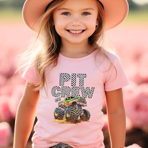 May include: Pink t-shirt with the words "PIT CREW" in a checkered pattern and a colorful monster truck graphic. The shirt is worn by a young person wearing a pink hat.