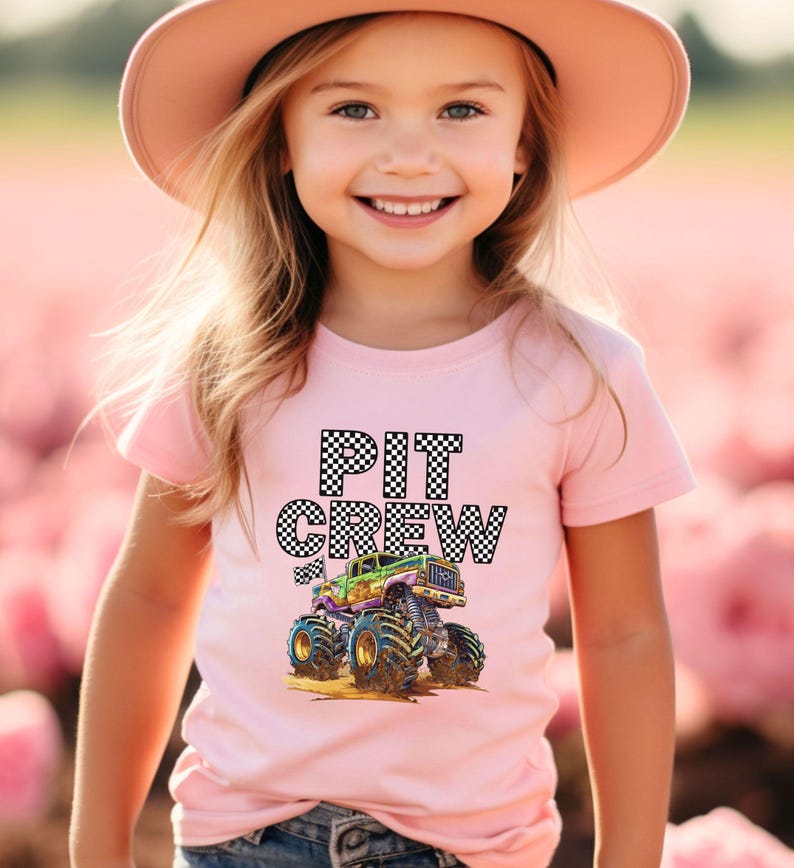 May include: Pink t-shirt with the words "PIT CREW" in a checkered pattern and a colorful monster truck graphic. The shirt is worn by a young person wearing a pink hat.