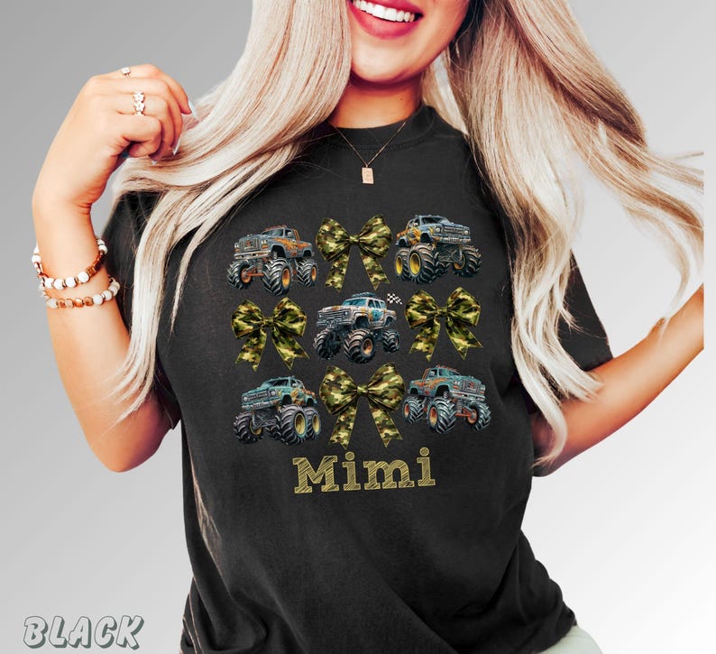 Woman wearing a black t-shirt featuring a design of monster trucks and camouflage bows. The word "Mimi" is printed in gold lettering below the trucks. The shirt is a comfortable, casual style.