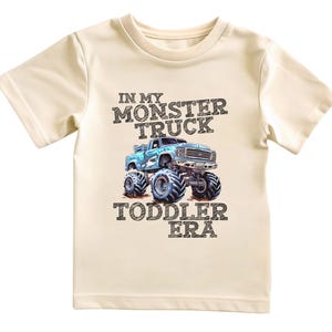 Puede incluir: Una camiseta beige claro para ni&ntilde;os peque&ntilde;os con un gr&aacute;fico de cami&oacute;n monstruo azul y el texto "In My Monster Truck Toddler Era".