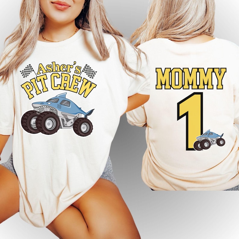 Pictured is a woman wearing a natural colored t-shirt which says "Asher's PIT CREW" custom name in yellow letters. there is a blue megalodon monster truck with 2 checkered flags. On the back of this 2-sided front back design is custom Name "MOMMY" in varsity letters font a big number 1 both in yellow and a smaller version of the truck