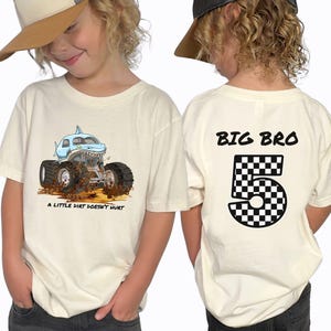a boy wearing a natural beige colored t-shirt showing front and back print . the front has a megalodon shark monster truck that says a "little dirt doesn't hurt" with a dirt floor and mud and dirt flying around it. The back of the shirt is a personalized name in black that says "Big Bro" and a checkered number 5. Any Name and Age can be applied. Adult , child, toddler and infant sizes.