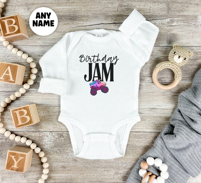 May include: White baby bodysuit with long sleeves. The text "Birthday JAM" is printed in black, with a colorful monster truck graphic. A wooden toy and alphabet blocks are in the background.