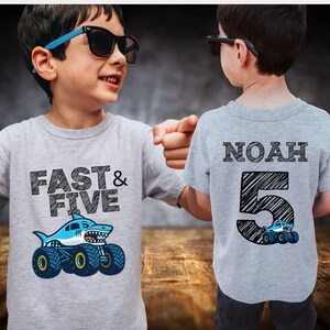 Pictured is a five year old boy wearing a light gray t-shirt. Front side has the text "FAST & FIVE" with a big shark monster truck graphic. The other shirt has the name "NOAH" and the number "5" with a monster truck graphic.