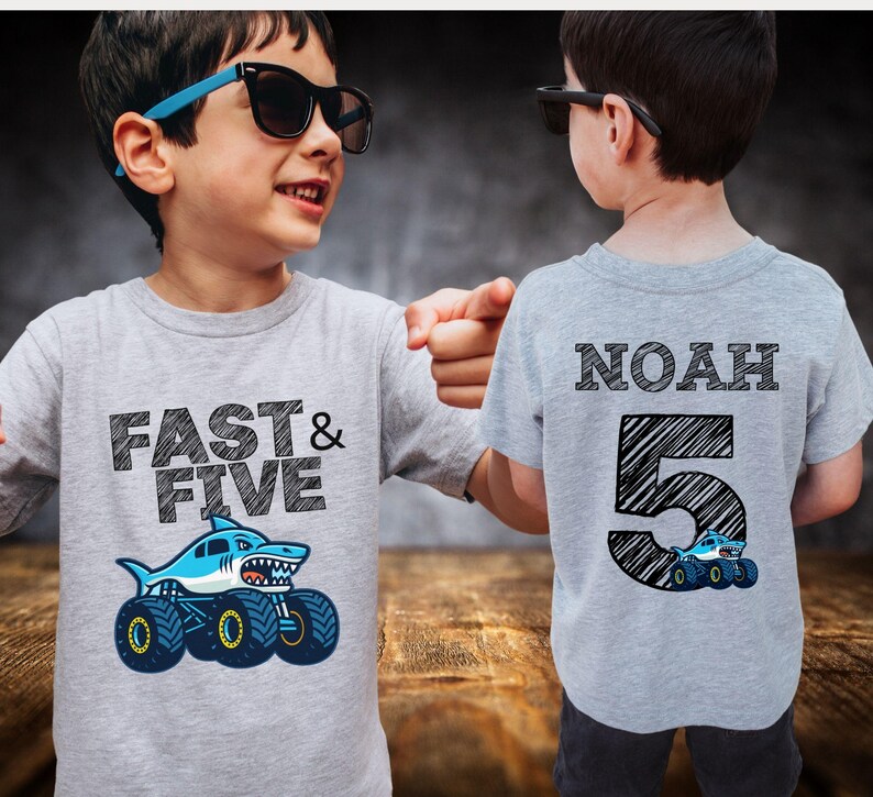 Pictured is a five year old boy wearing a light gray t-shirt. Front side has the text "FAST & FIVE" with a big shark monster truck graphic. The other shirt has the name "NOAH" and the number "5" with a monster truck graphic.