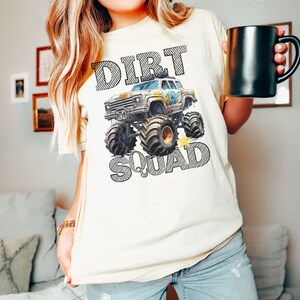 Puede incluir: Camiseta de color marfil con un gr&aacute;fico de monster truck y las palabras "DIRT SQUAD" en negrita y con contorno. El monster truck tiene detalles en azul y amarillo. La camiseta es de corte holgado.