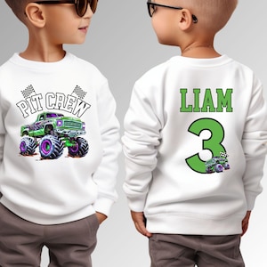 Op de afbeelding: Witte sweatshirt met een groene monstertruck-afbeelding en de tekst "PIT CREW". De achterkant van de sweatshirt heeft de naam "LIAM" en het nummer "3" in groen. Een kleine monstertruck-afbeelding staat onder het nummer.