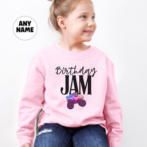 May include: A light pink sweatshirt with the words "Birthday JAM" in black script and block letters. Below the text is a colorful monster truck graphic. A white circle with the words "ANY NAME" is in the upper left corner.
