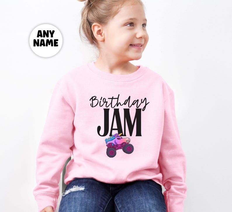 May include: A light pink sweatshirt with the words "Birthday JAM" in black script and block letters. Below the text is a colorful monster truck graphic. A white circle with the words "ANY NAME" is in the upper left corner.