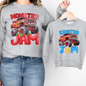 May include: Two gray sweatshirts with matching designs. One sweatshirt features the text "MOMSTER JAM" with a red monster truck graphic. The other sweatshirt has the text "KIDSTER JAM" with a similar truck graphic. Both sweatshirts are long-sleeved.