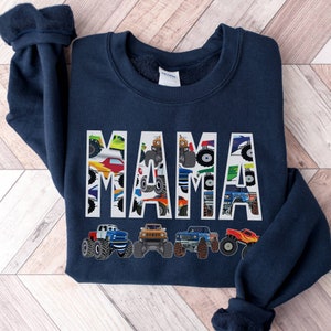 Pictured is a Navy Blue Sweatshirt Which has the Custom Name MAMA in large letters with monster trucks in the letters and outlined in white. Underneath are 4 monster truck graphics
