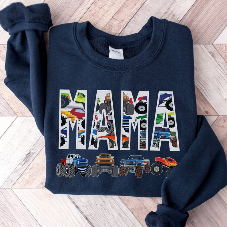 Pictured is a Navy Blue Sweatshirt Which has the Custom Name MAMA in large letters with monster trucks in the letters and outlined in white. Underneath are 4 monster truck graphics