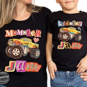 Puede incluir: Camisetas negras con gr&aacute;ficos de monster trucks. Una camiseta dice "Momster Jam" y la otra "Kidster Jam". Los monster trucks son amarillos con neum&aacute;ticos grandes. Las camisetas tienen texto colorido y un coraz&oacute;n a cuadros.