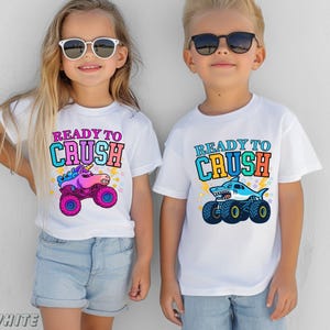 May include: Two children wearing white t-shirts with the text "READY TO CRUSH". One shirt features a unicorn monster truck, the other a shark monster truck. Both shirts have colorful graphics and are paired with denim shorts.