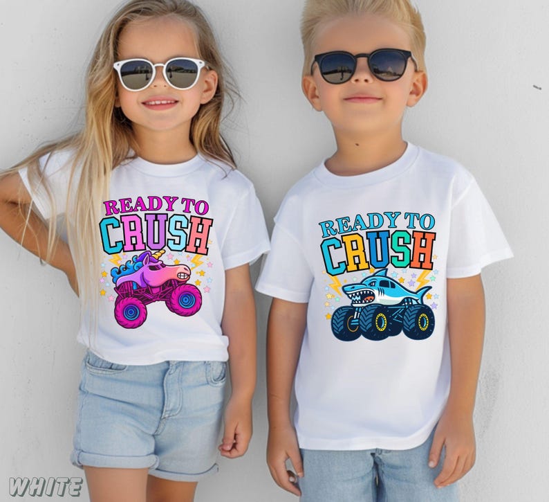 May include: Two children wearing white t-shirts with the text "READY TO CRUSH". One shirt features a unicorn monster truck, the other a shark monster truck. Both shirts have colorful graphics and are paired with denim shorts.