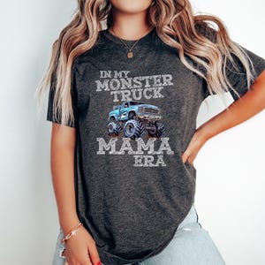 Puede incluir: Una camiseta gris oscuro con un gr&aacute;fico de cami&oacute;n monstruo azul y el texto "In My Monster Truck Mama Era".