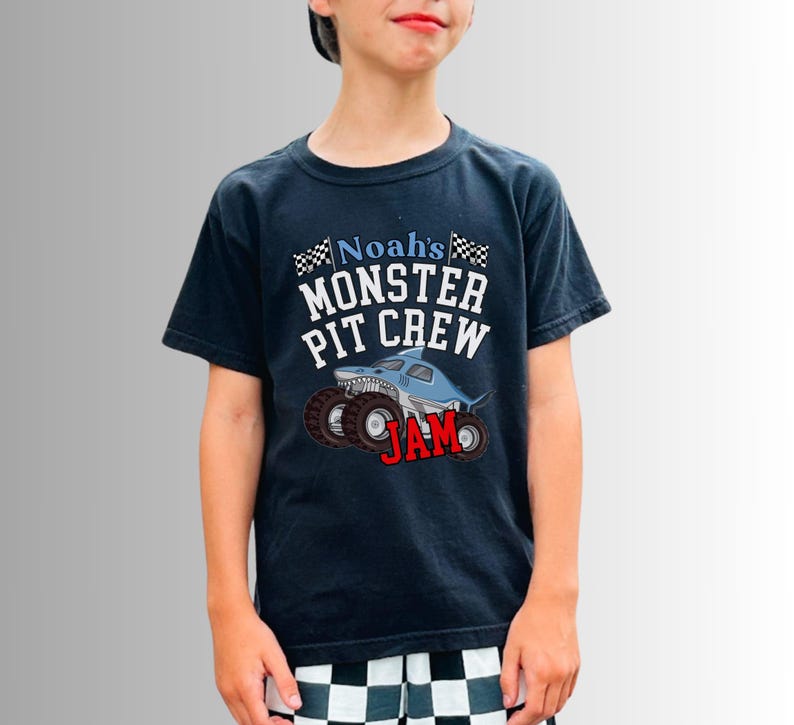 Pictured is a boy wearing a navy blue t-shirt with the text "Noah's Monster Pit Crew JAM" and a graphic of a shark-shaped monster truck. The shirt has a crew neck and short sleeves. The child is wearing a black baseball cap.
