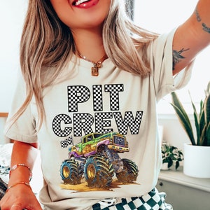 Puede incluir: Una camiseta blanca con un diseño a cuadros blanco y negro que dice "PIT CREW" con un gráfico de camión monstruo debajo del texto.