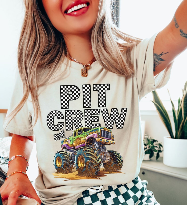 May include: Beige t-shirt with the words "PIT CREW" in a black and white checkered pattern. Below the text is a colorful monster truck graphic with large tires and mud. The shirt is a casual style.