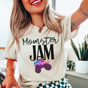 May include: Cream-colored t-shirt with the text "Momster JAM" in black script and block letters. A colorful monster truck with a unicorn horn is featured below the text. The truck has purple wheels and a pink and blue body.
