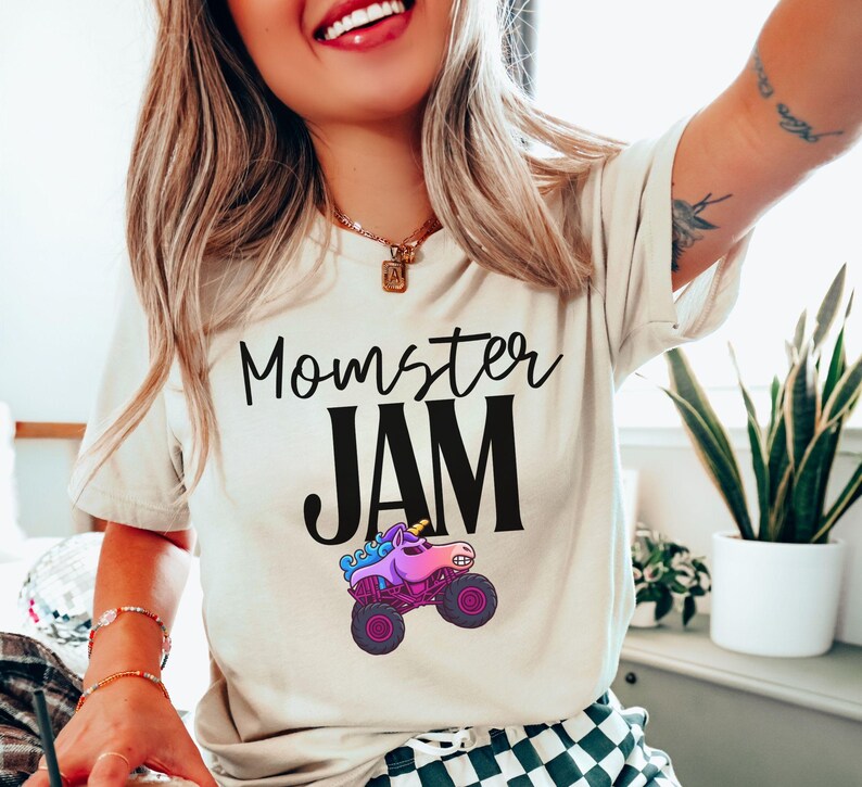May include: Cream-colored t-shirt with the text "Momster JAM" in black script and block letters. A colorful monster truck with a unicorn horn is featured below the text. The truck has purple wheels and a pink and blue body.