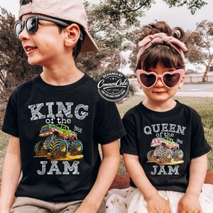 May include: Two children wearing black t-shirts with monster truck graphics. One shirt says "KING of the JAM" and the other says "QUEEN of the JAM". The shirts have colourful monster truck designs and the text is in white. The children are wearing sunglasses.