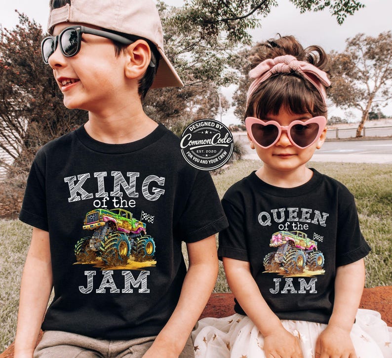 May include: Two children wearing black t-shirts with monster truck graphics. One shirt says "KING of the JAM" and the other says "QUEEN of the JAM". The shirts have colourful monster truck designs and the text is in white. The children are wearing sunglasses.
