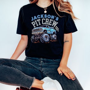 Pictured is a woman wearing a Black t-shirt featuring a blue shark monster truck graphic with the text "Jackson's Pit Crew". The truck has large tires and a checkered flag.