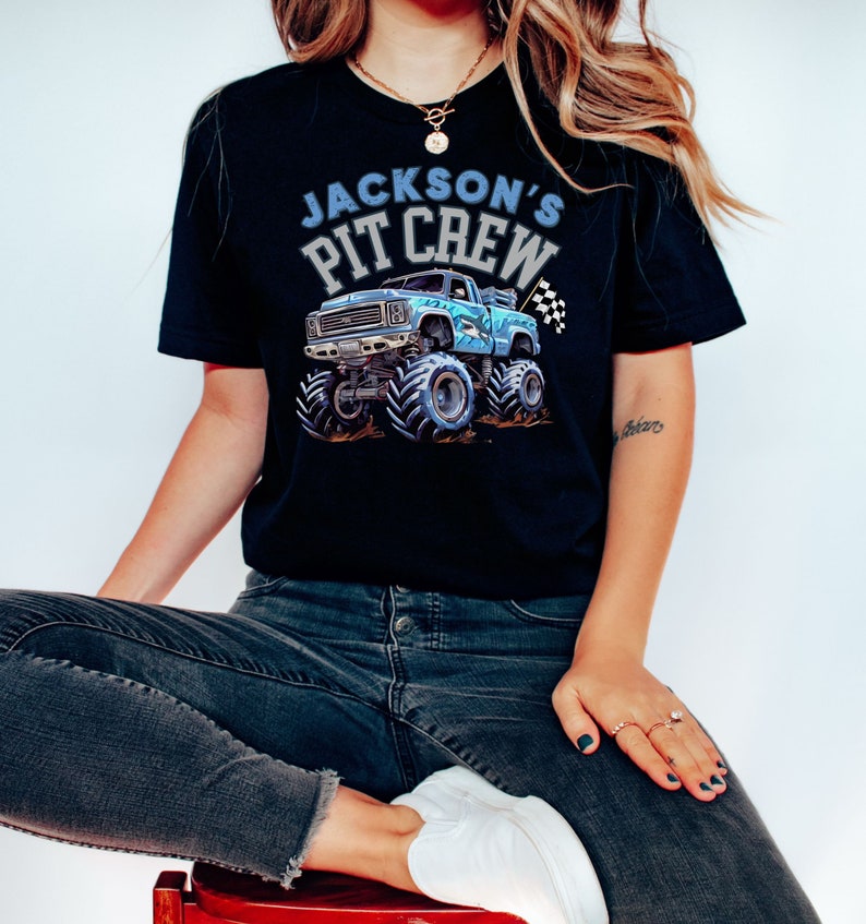 Pictured is a woman wearing a Black t-shirt featuring a blue shark monster truck graphic with the text "Jackson's Pit Crew". The truck has large tires and a checkered flag.