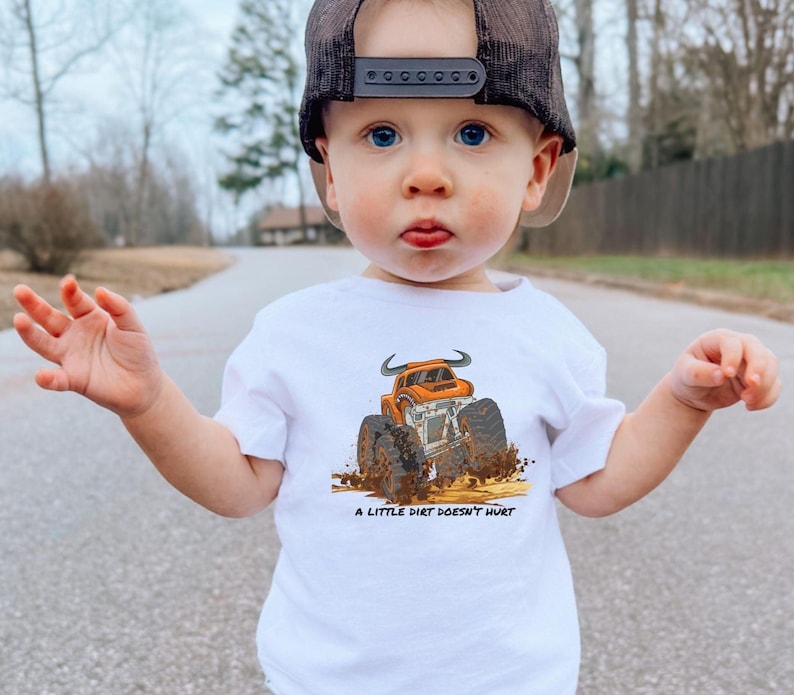 Cute boy wearing a white tee with an orange bull monster truck with the text "A little dirt doesn't hurt" underneath the graphic