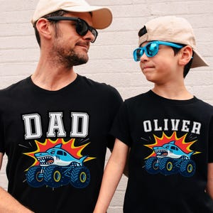 Dad and Son are wearing black t-shirts with matching shark monster truck designs. One shirt says "DAD" in white block letters, the other says "OLIVER". Both shirts feature a blue shark on a monster truck with a red and yellow burst behind the graphic.