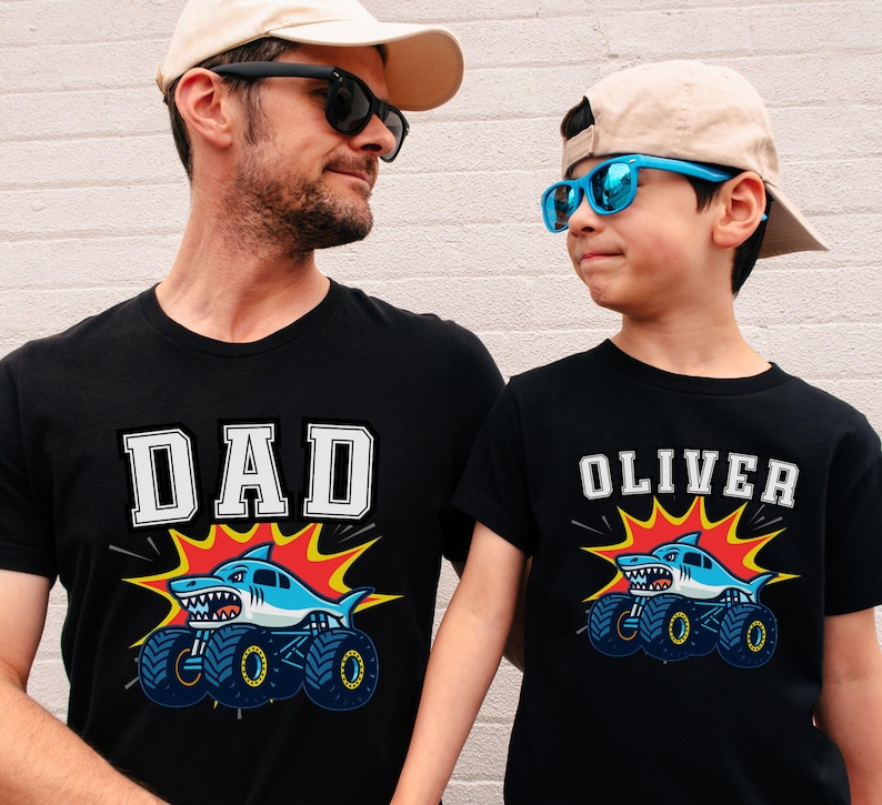 Dad and Son are wearing black t-shirts with matching shark monster truck designs. One shirt says "DAD" in white block letters, the other says "OLIVER". Both shirts feature a blue shark on a monster truck with a red and yellow burst behind the graphic.