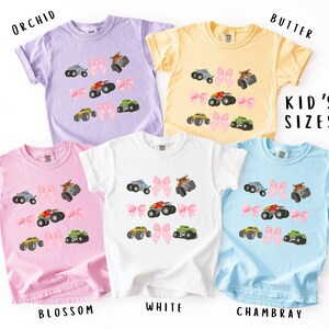 K&ouml;nnte beinhalten: Eine Auswahl an Kinder-T-Shirts in den Farben Orchidee, Butter, Blossom, Wei&szlig; und Chambray. Jedes Shirt zeigt ein Monster-Truck-Design mit einer rosa Schleife. Der Text "KID'S SIZES" ist ebenfalls sichtbar.